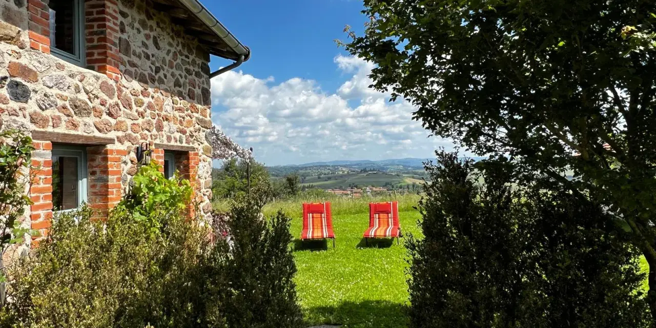 Maison en pierre avec vue panoramique sur les collines, deux chaises longues rouges invitent à la détente. Parfait pour découvrir les destinations Rhône-Alpes.