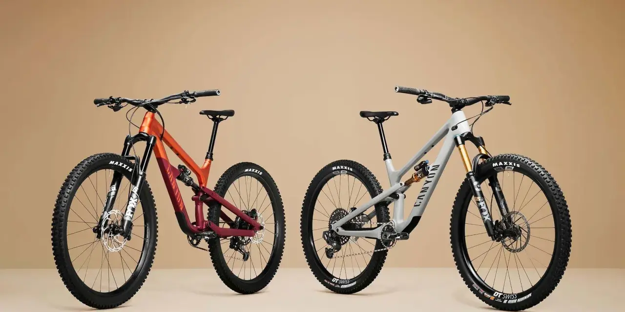 Two full-suspension mountain bikes, one orange and maroon, the other grey, are displayed side-by-side, showcasing the 27.5 vs 29 wheel size debate.