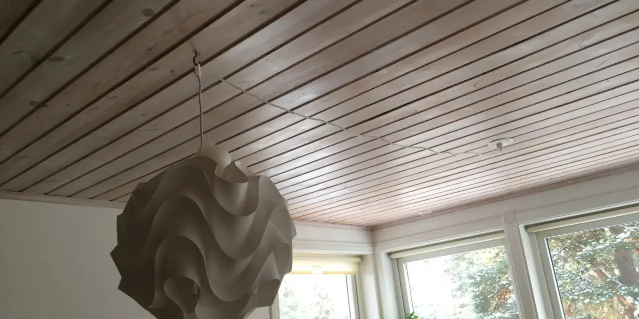 En vit lampskärm hänger från ett tak av träpanel. En tunn sladd dras i taket för att ansluta lampan.