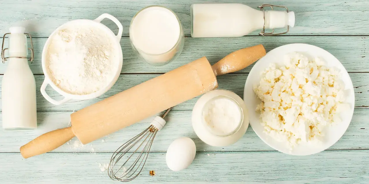 Zutaten für butter ersatz backen: Mehl, Milch, Quark, Ei, Schneebesen und Nudelholz auf blauem Holzuntergrund.