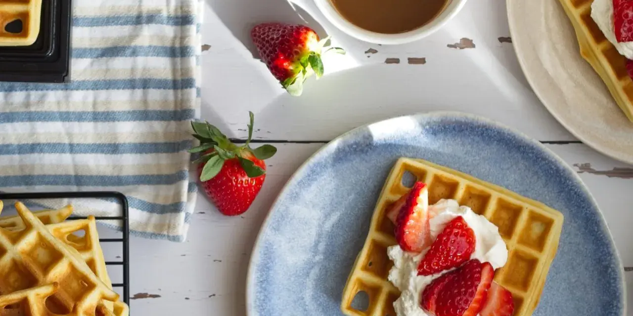 Frische belgische Waffeln mit Erdbeeren und Sahne auf einem blauen Teller, daneben eine Tasse Kaffee und weitere Waffeln auf einem Gitter.