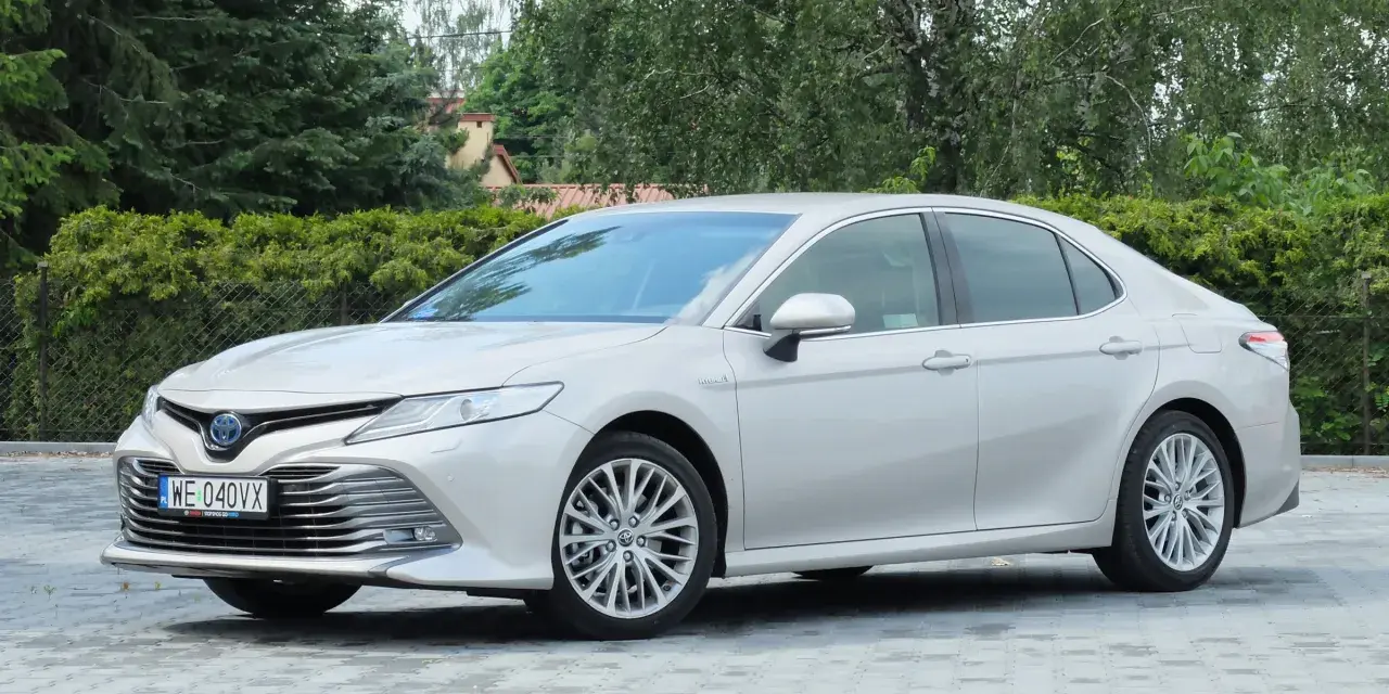 Srebrna Toyota Camry 2.5 Hybrid stoi na wybrukowanym podjeździe, otoczona zielenią.