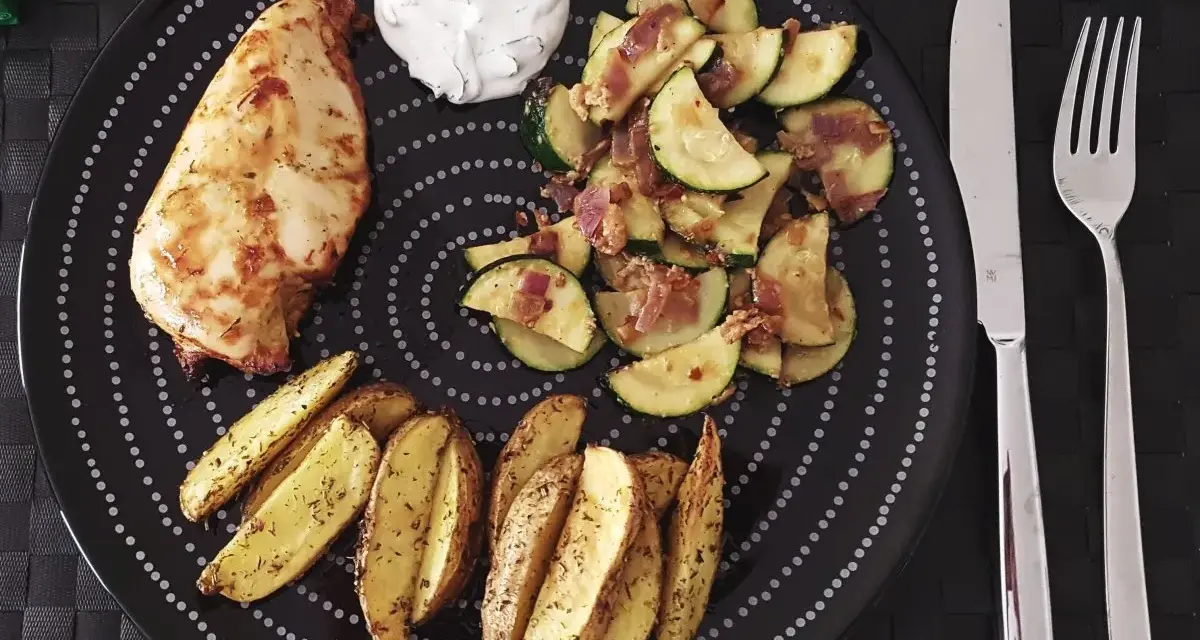 Ein Teller mit Hähnchenbrust, Kartoffelspalten und Zucchinigemüse, perfekt für Airfryer Rezepte. Dazu ein Klecks Dip.