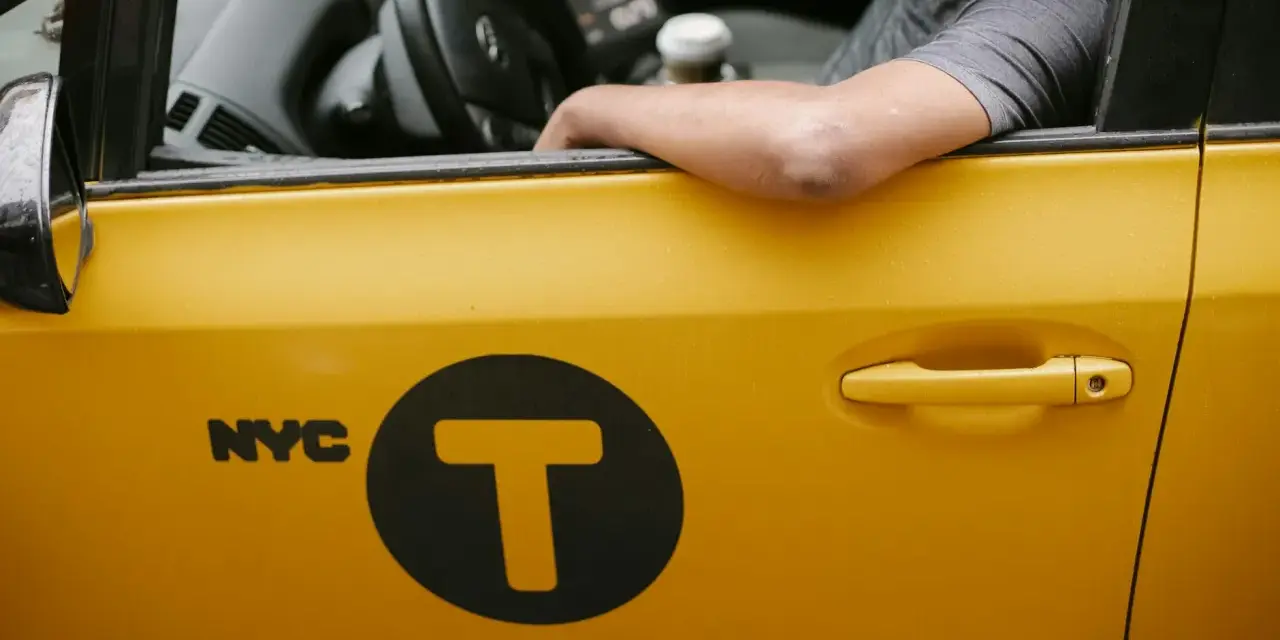 Kierowca żółtej taksówki NYC z logo "T" w kole. Zastanawiasz się, ile zarabia taksówkarz w tym mieście?