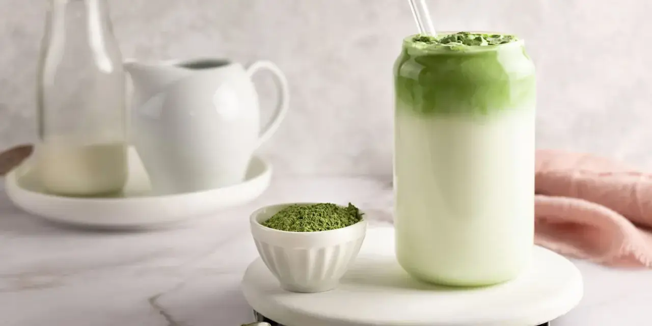Pyszny matcha latte w szklance, obok miseczka z zielonym proszkiem i łyżeczka.