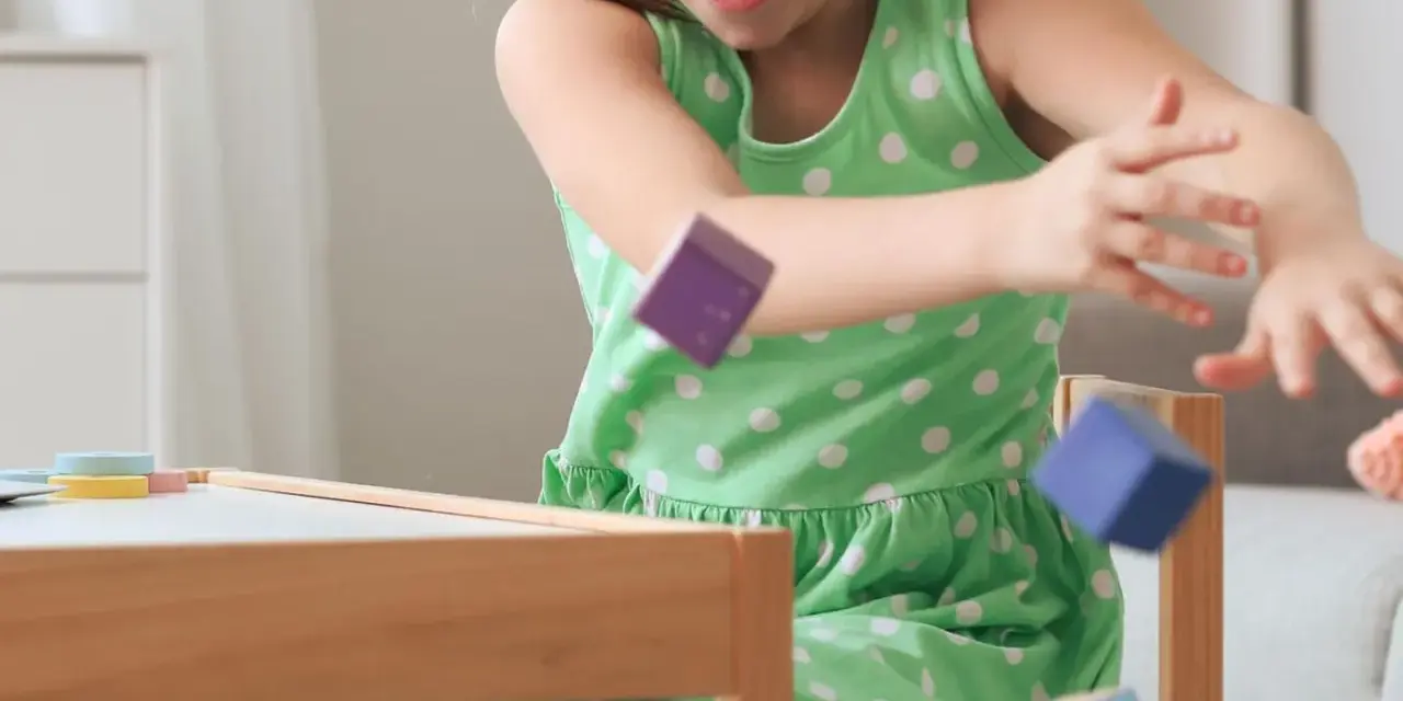 Ein Mädchen in einem grünen Kleid wirft bunte Holzklötze. Ein Versuch, laute Kinder zur Ruhe zu bringen, indem man sie zum Spielen anregt.