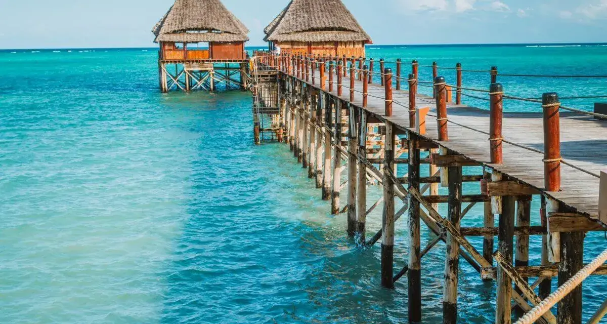 Drewniany pomost prowadzi do chat na palach nad turkusową wodą. Zanzibar, pora deszczowa, ale słońce świeci.