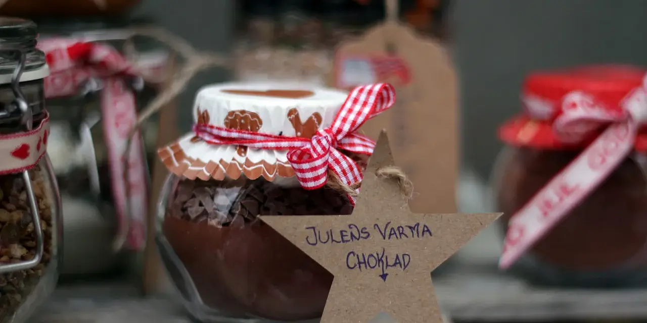 Burkar med ingredienser för varm choklad jul, dekorerade med röda band och pepparkaksfigurer. En stjärna med texten "Julens varma choklad" hänger framför.