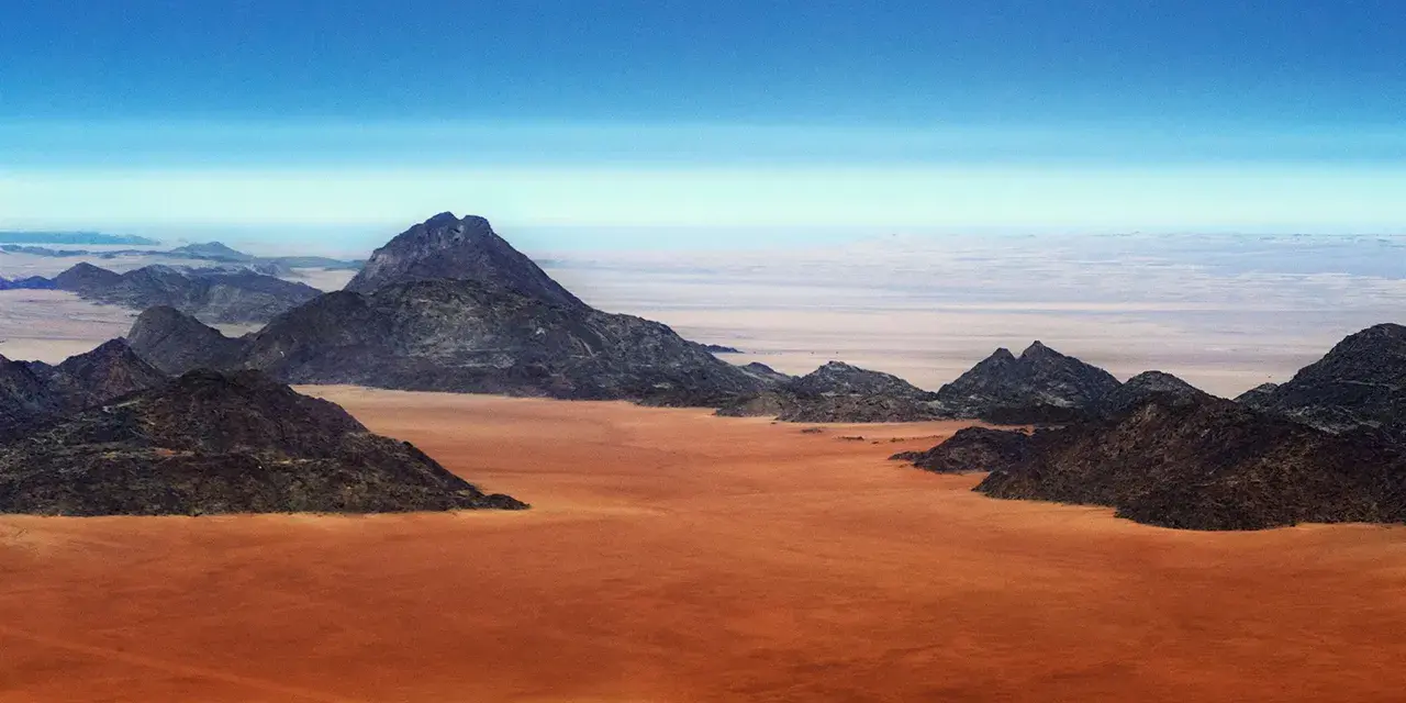Pustynny krajobraz w **Afryce, Namibia**. Czerwona ziemia i skaliste góry pod błękitnym niebem.