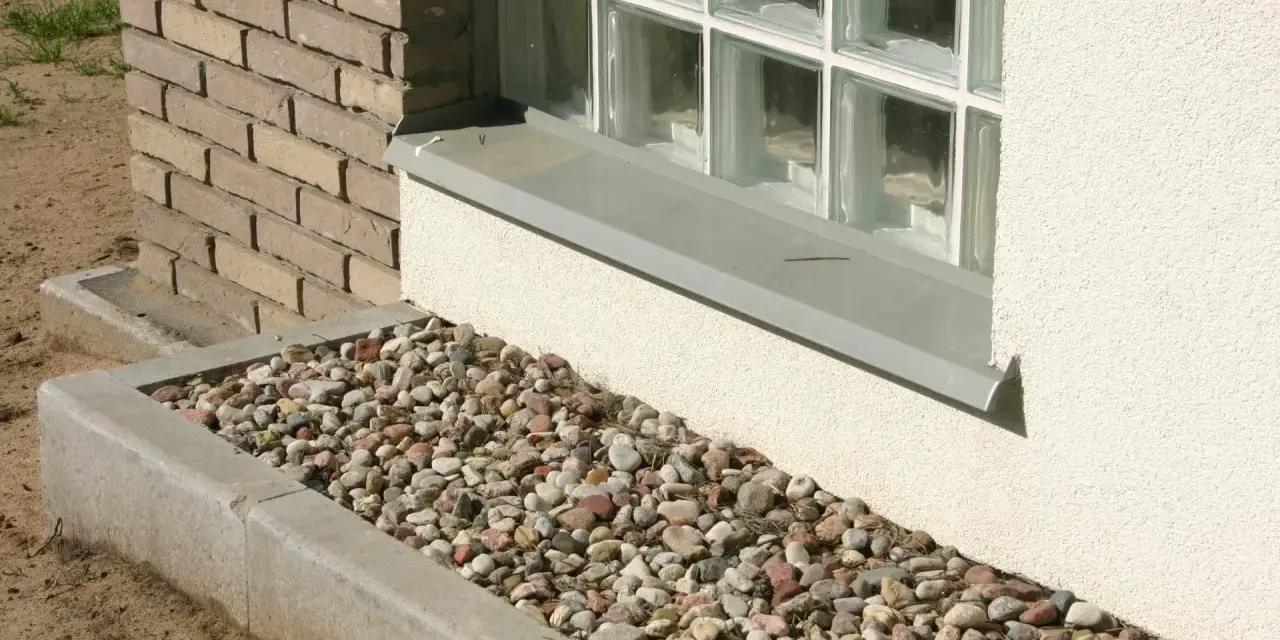 Kamienna opaska żwirowa wokół domu, otaczająca betonowy murek i okno z bloczków szklanych.