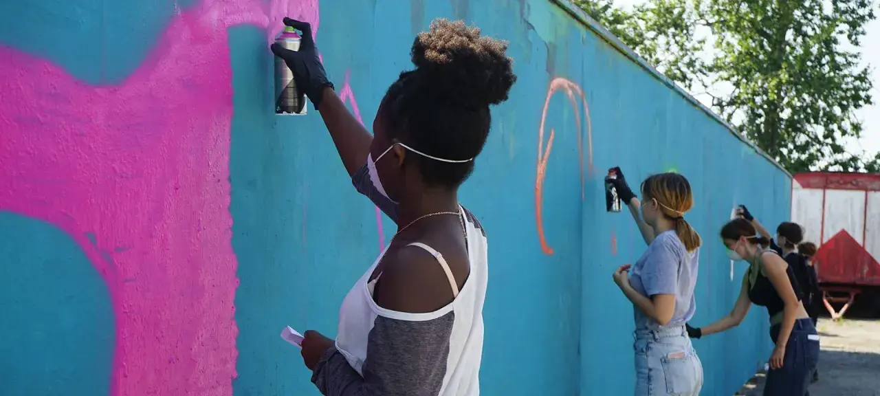 Ungdomar målar graffiti på en blå vägg. En flicka med afrohår sprayar rosa färg.