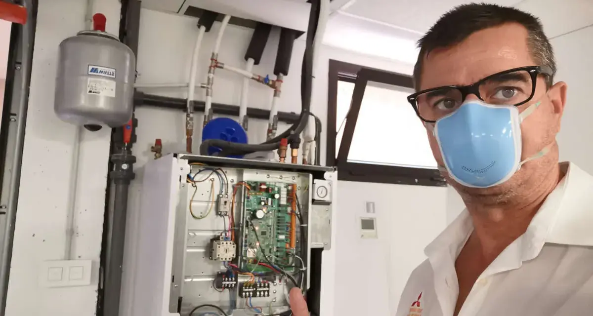 Técnico con mascarilla realiza mantenimiento aerotermia. Equipo abierto, circuito visible. Pulgar arriba.