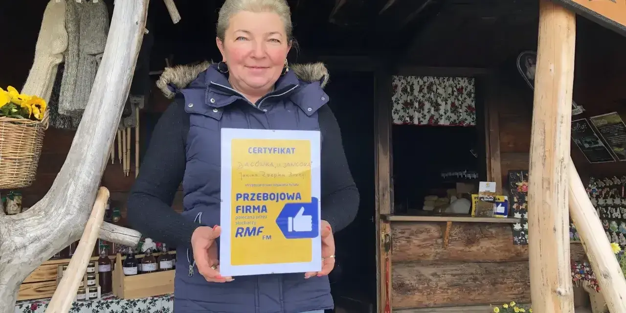 Kobieta z certyfikatem "Przejściowa Firma" przed bacówką. W tle widać rękodzieło i przetwory, a także tablicę z napisem "Bacówka u Jancora".