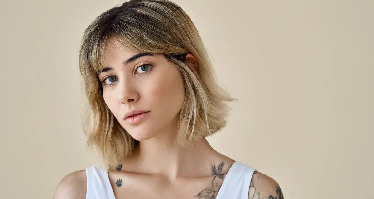Kobieta z tatuażami na ramionach i dekolcie, z modną fryzurą long bob i grzywką.
