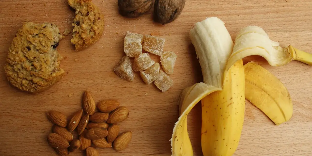 Na drewnianym blacie leżą orzechy, migdały, kawałek ciastka, kandyzowany imbir i banan. To świetne węglowodany złożone produkty.