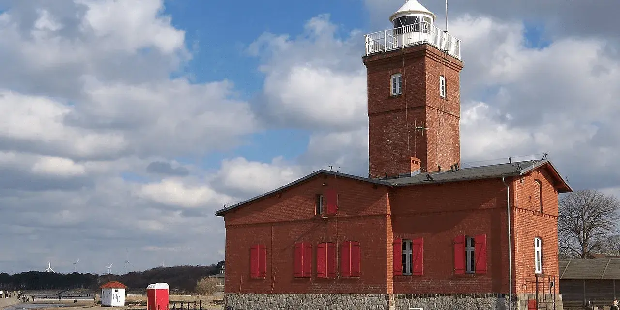 Ceglana latarnia morska Darłowo z czerwonymi okiennicami, na tle błękitnego nieba z chmurami.