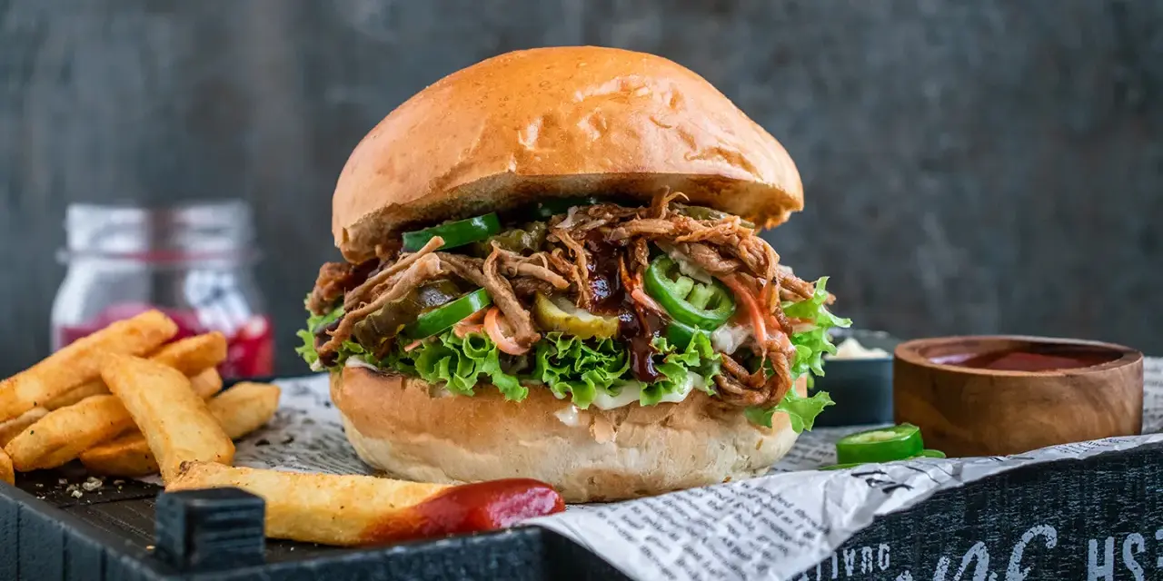 Saftiger pulled pork burger mit Salat, Jalapeños und Gurken. Die Zutaten für diesen Burger sind perfekt aufeinander abgestimmt. Dazu Pommes und Dip.