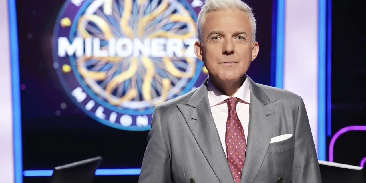 Hubert Urbański w garniturze, na tle koła z napisem "Milionerzy". Co dajemy komuś na drogę milionerzy? Wiedzę i odwagę!