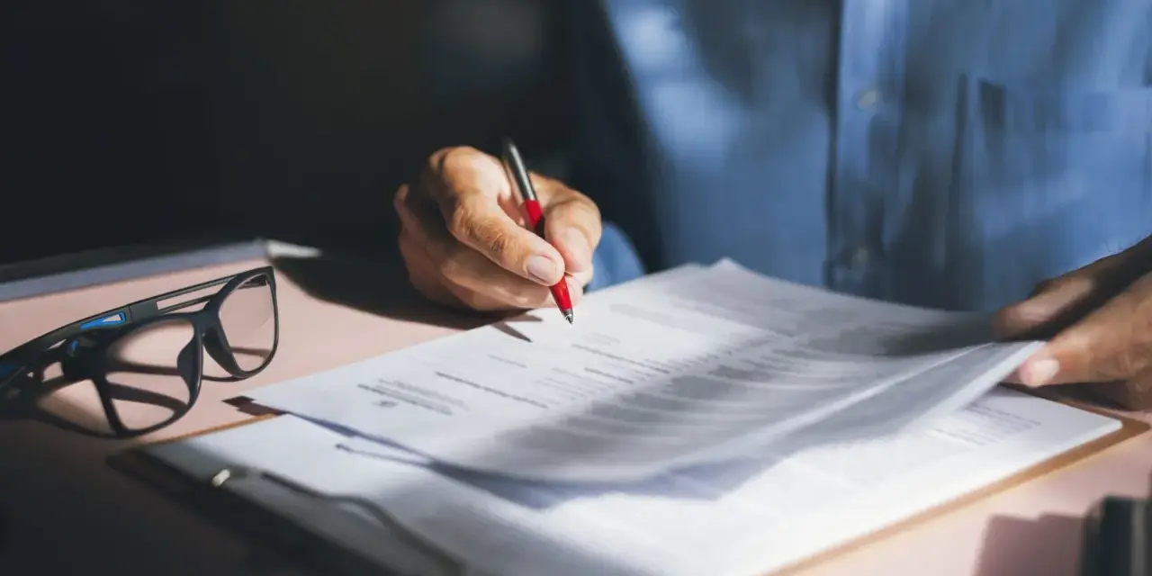 Persona revisa y firma una nota de encargo inmobiliaria, con gafas y bolígrafo rojo sobre documentos.
