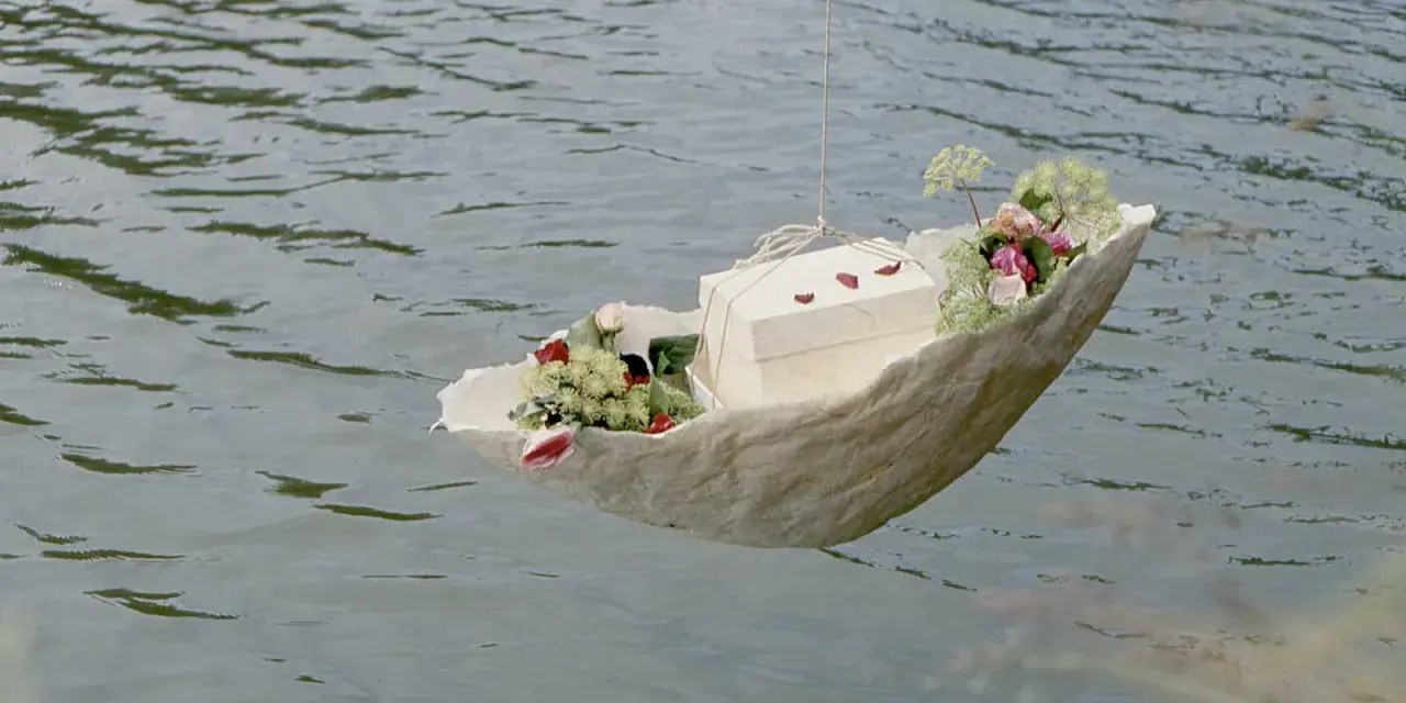 Ein Schiffchen mit Blumen und einer Schachtel schwimmt auf dem Wasser. Eine würdevolle Idee für Kinderbeerdigungen.