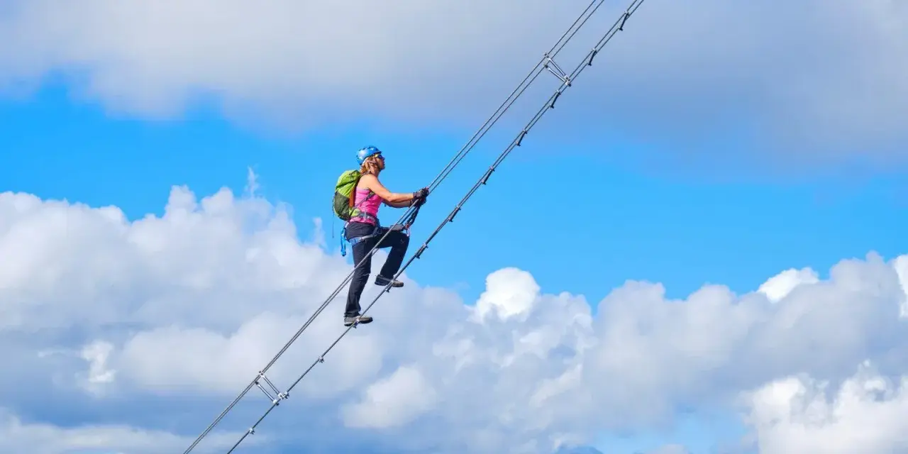 Frau klettert auf dem Donnerkogel Klettersteig über eine Hängebrücke, umgeben von blauem Himmel und Wolken.
