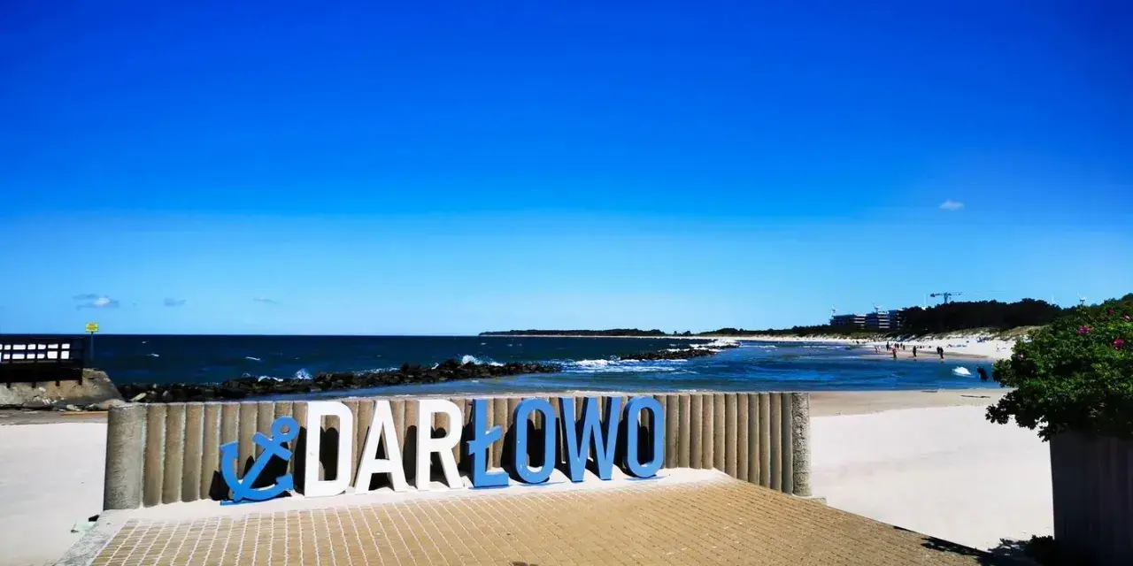Nadmorskie miasto nad Wieprzą, Darłowo, wita turystów na piaszczystej plaży. Błękitne niebo i morze tworzą idealne tło dla napisu.