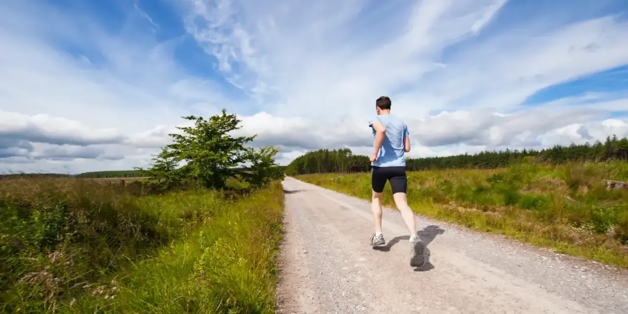 Uomo corre su strada sterrata, ideale per la preparazione 10 km principianti. Cielo azzurro con nuvole bianche.