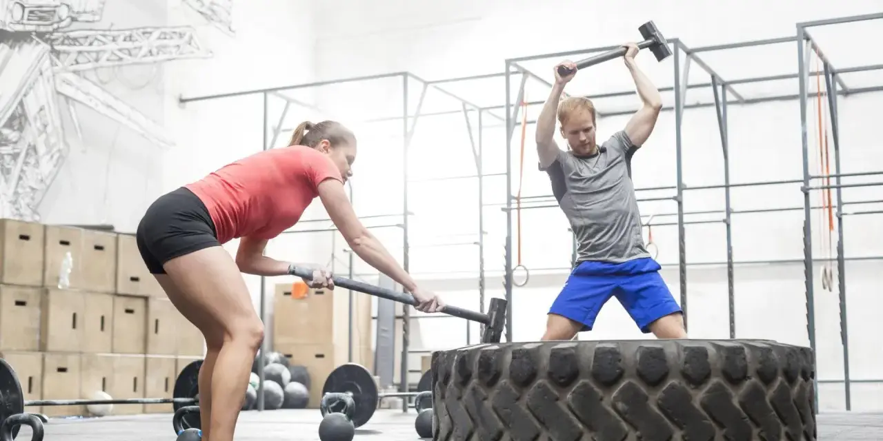 Intensywny trening co to jest crossfit: mężczyzna i kobieta uderzają młotami w wielką oponę w siłowni.