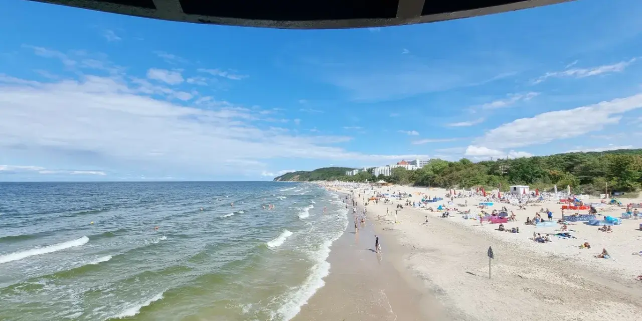 Słoneczna plaża z tłumem turystów, gdzie jest najcieplejsze morze. Woda faluje, a na piasku rozłożone są parawany i ręczniki.