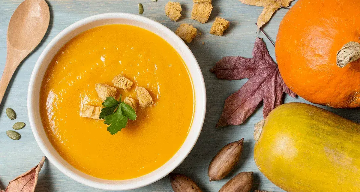 Un reconfortante tazón de puré de calabaza, adornado con picatostes y perejil, junto a calabazas, hojas de otoño y nueces.
