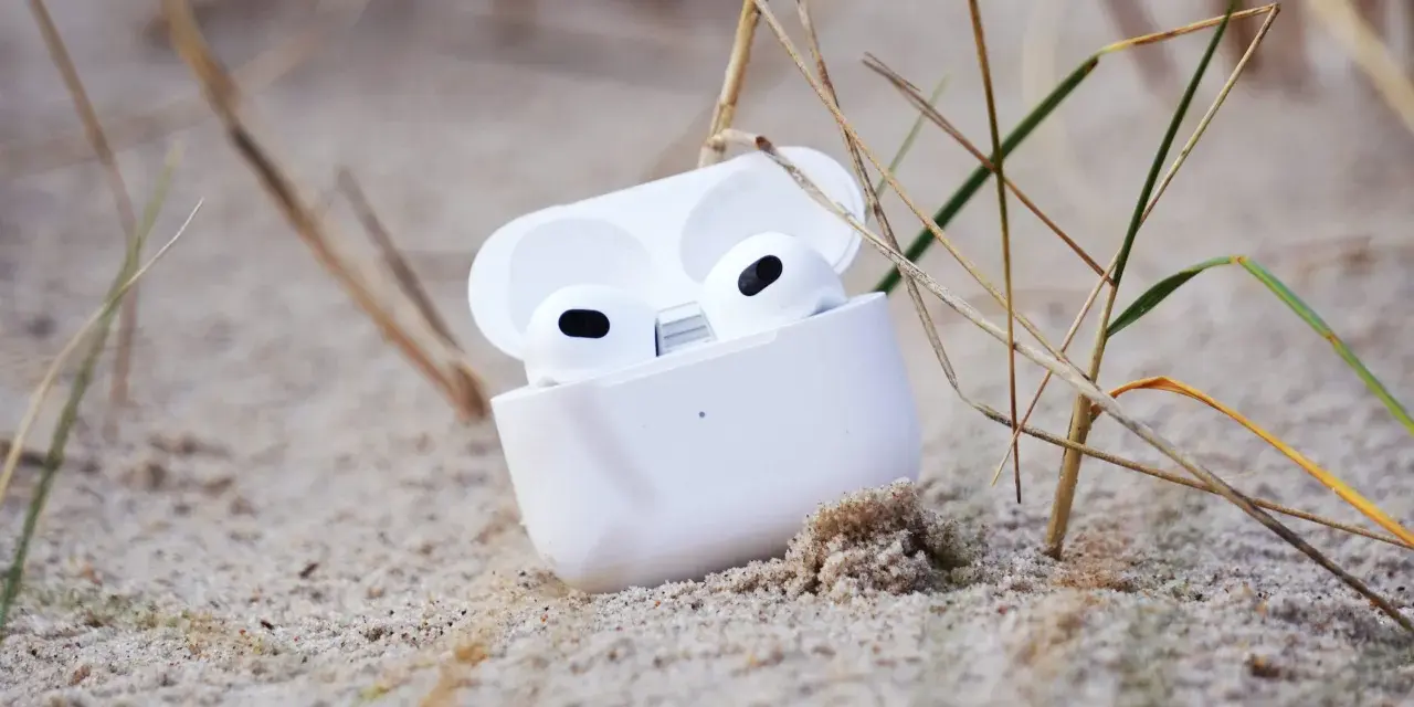 Weiße AirPods im geöffneten Ladecase liegen im Sand, umgeben von Gräsern.
