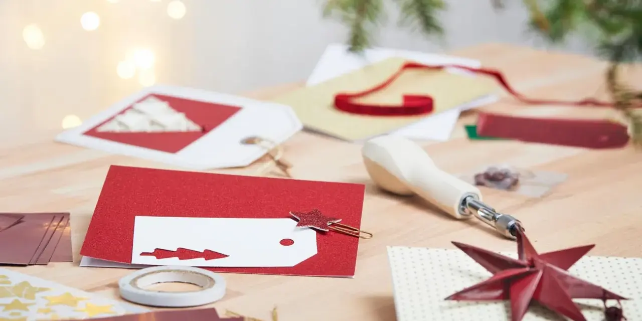 Förberedelser för att måla julkort. Rött glitterpapper, vita etiketter med julgransmotiv och en stjärnformad gemklämma ligger på ett träbord.