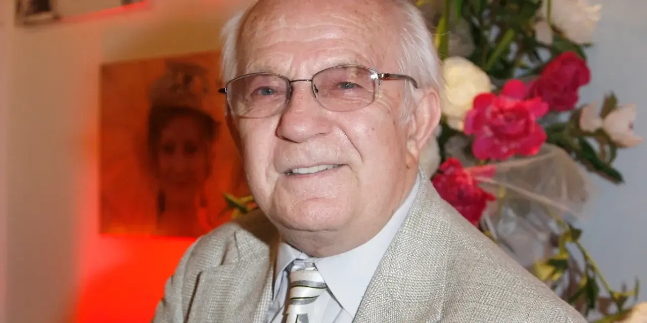 Uśmiechnięty Jerzy Antczak, reżyser znany z niezapomnianych filmów, w eleganckim garniturze, na tle bukietu kwiatów i portretu.