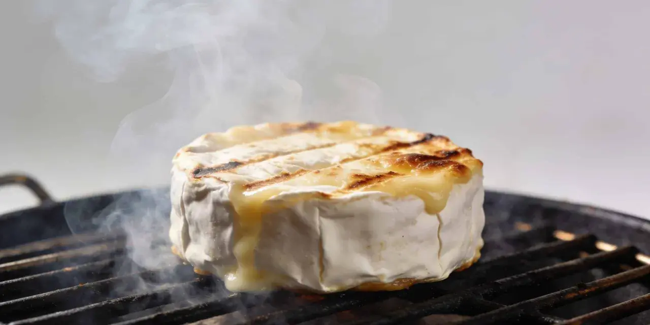 Un camembert au barbecue, fondant et fumant, cuit sur une grille. La croûte blanche se craquelle, révélant un cœur doré et coulant.