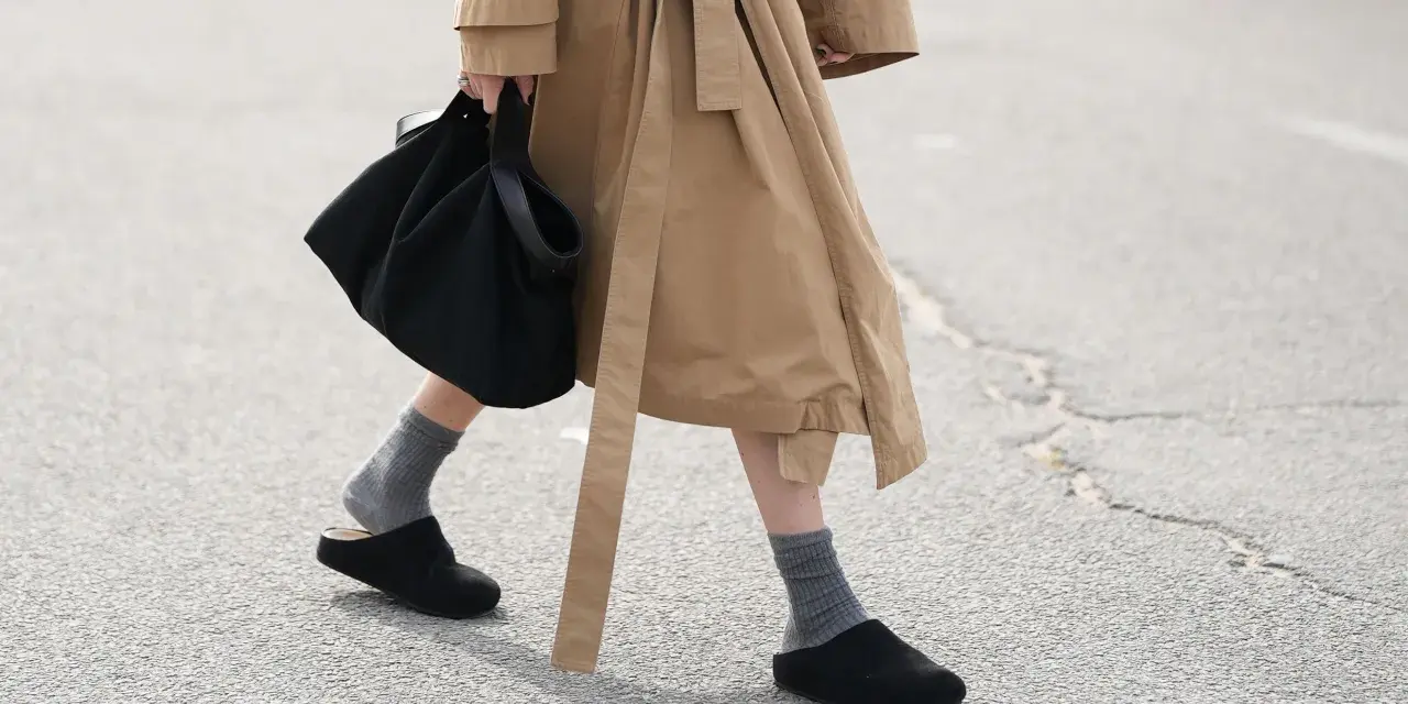 Persona camina con zuecos negros y calcetines grises, llevando un bolso negro y un abrigo beige.