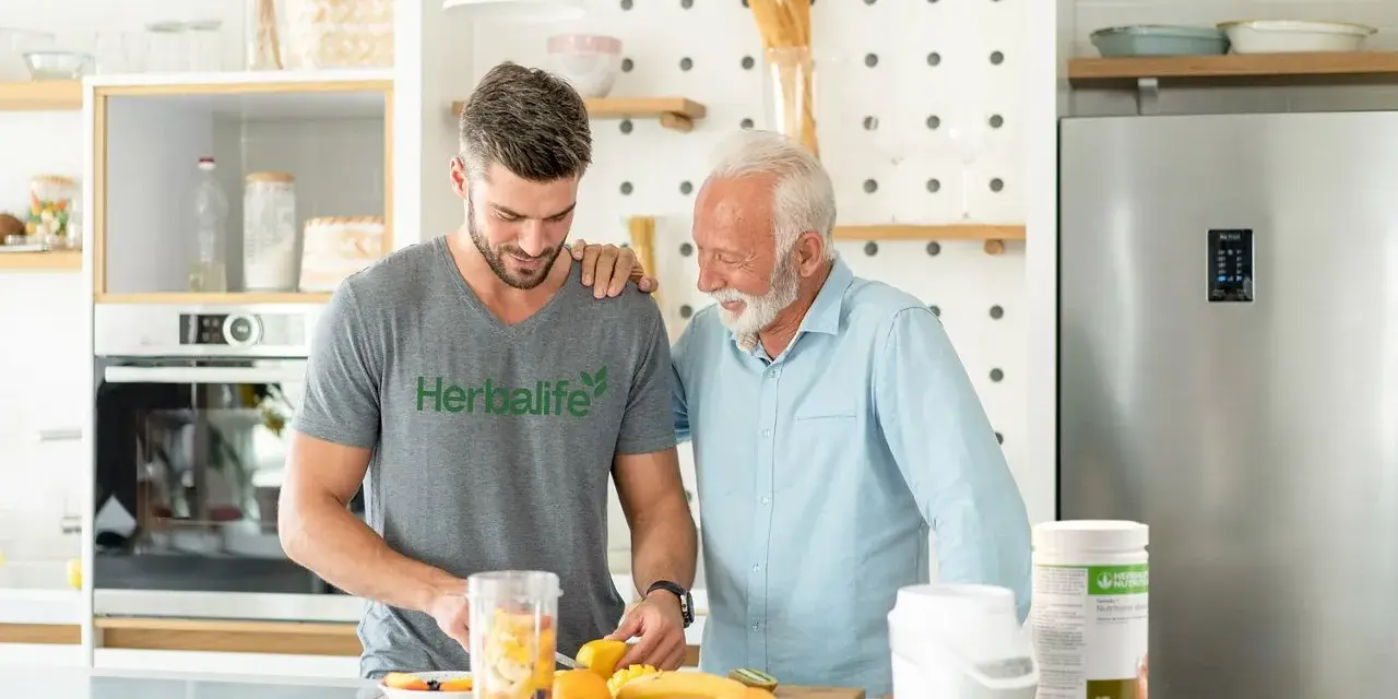 Två män i ett kök förbereder en smoothie med frukt. En burk med vegetariskt protein står på bänken.