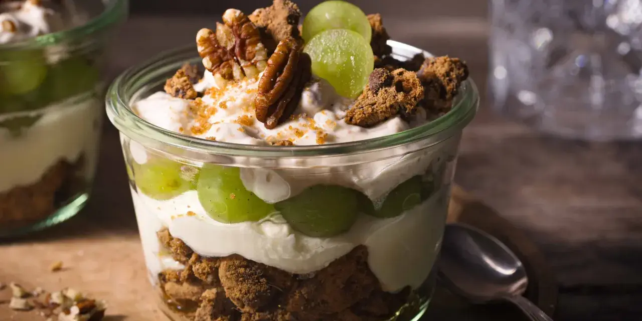 Ein Glas mit cremigem Dessert, geschichtet mit grünen Trauben, zerbröselten Keksen und Nüssen. Ein köstliches Trauben-Cookie-Dessert, perfekt für jeden Anlass.