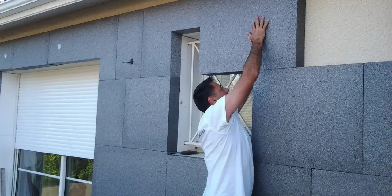 Un ouvrier pose des panneaux isolants gris pour l'isolation par l'extérieur d'une maison, près d'une fenêtre avec volet roulant.
