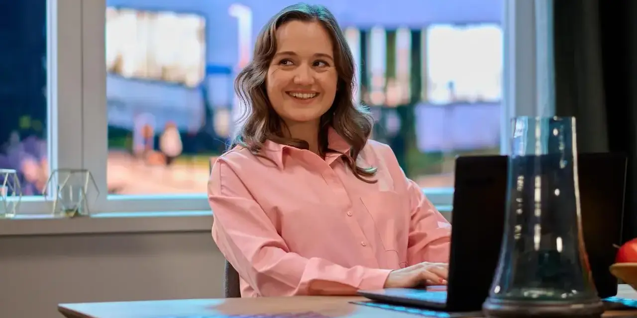 Uśmiechnięta Dorota Krempa pracuje przy laptopie, siedząc przy stole. W tle widać okno z nocnym widokiem.