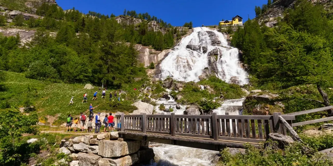 Cascata imponente con escursionisti su un sentiero, un ponte di legno attraversa un torrente. Un esempio di cosa vedere in Piemonte per gli amanti della natura.