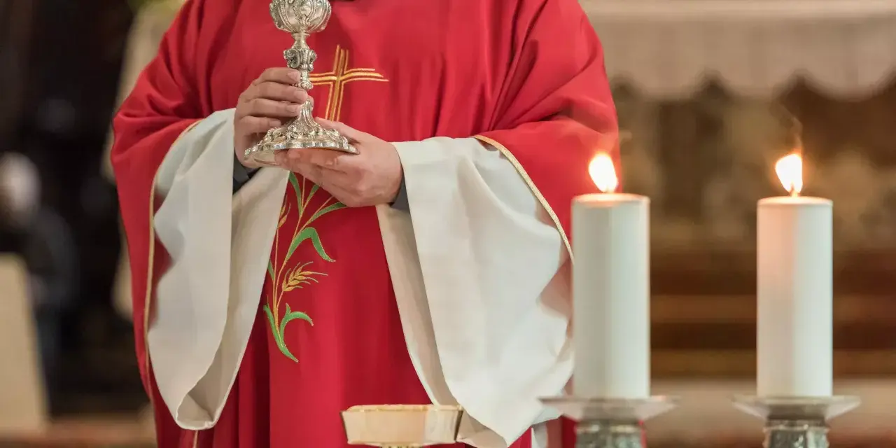 Kapłan w czerwonej szacie z krzyżem trzyma kielich podczas konsekracji. Płoną dwie świece.