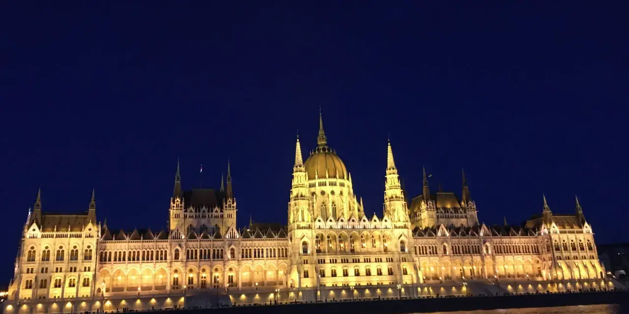 Budynek Parlamentu w Budapeszcie nocą, oświetlony, z odbiciem w Dunaju. Warto sprawdzić ceny na Węgrzech przed podróżą.