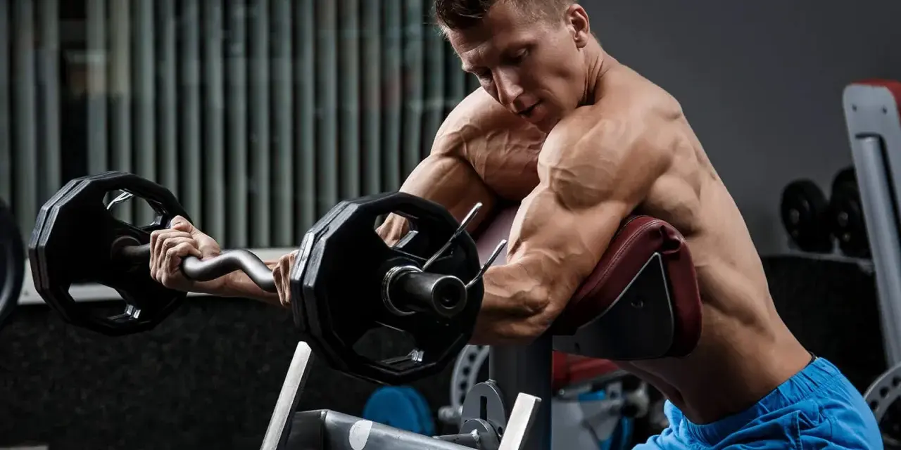 Mężczyzna wykonuje ćwiczenia na biceps i triceps z użyciem sztangi łamanej na modlitewniku.