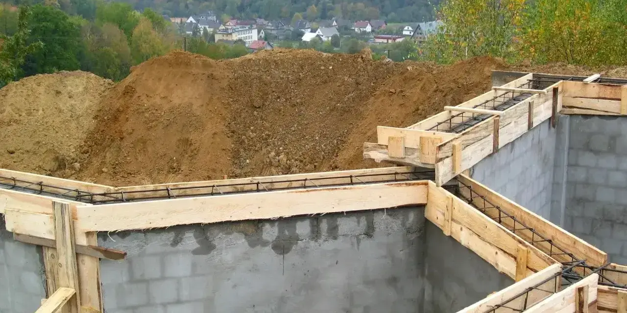 Fundamenty domu w budowie, zbrojenie i szalunki. Widać kopce ziemi i zabudowania w tle. Ważne jest pozwolenie na budowę, by wszystko szło zgodnie z planem.