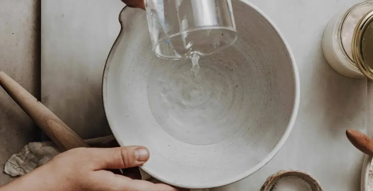 Wasser wird in eine Schüssel gegossen, vielleicht zu viel Wasser im Brotteig. Ein Nudelholz liegt daneben.