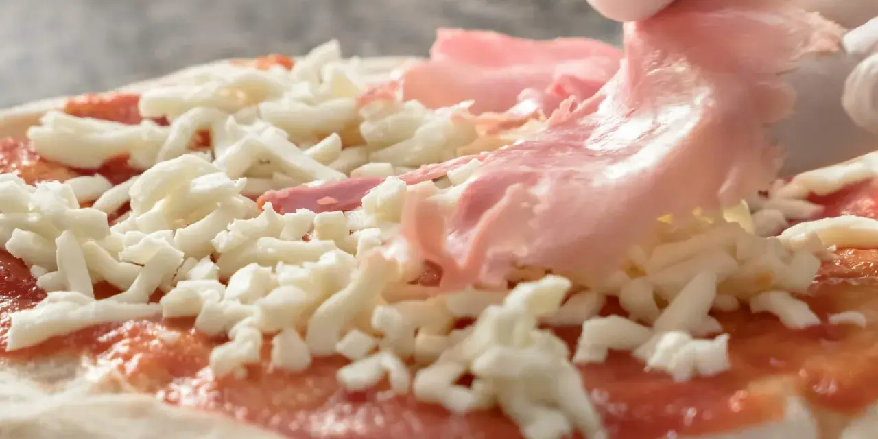 Ręka w białej rękawiczce kładzie plasterki szynki na pizzy z serem i sosem pomidorowym.