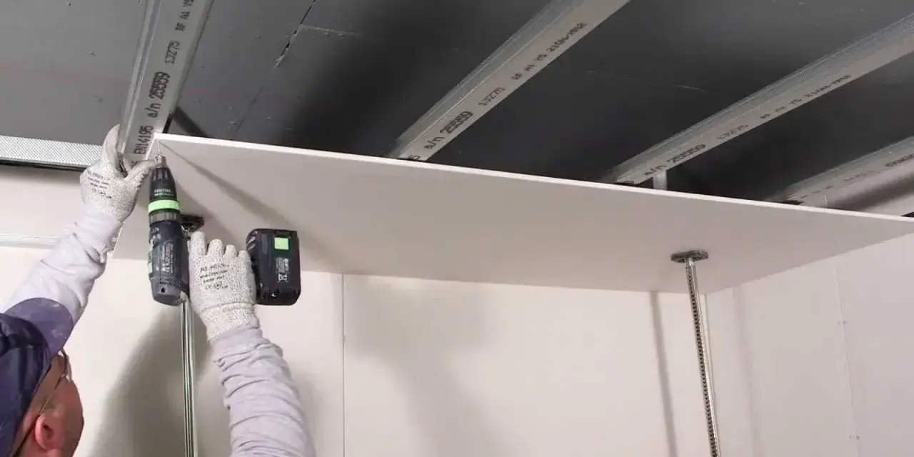 Persona con guanti avvita una lastra di cartongesso al soffitto metallico, un progetto di cartongesso fai da te.