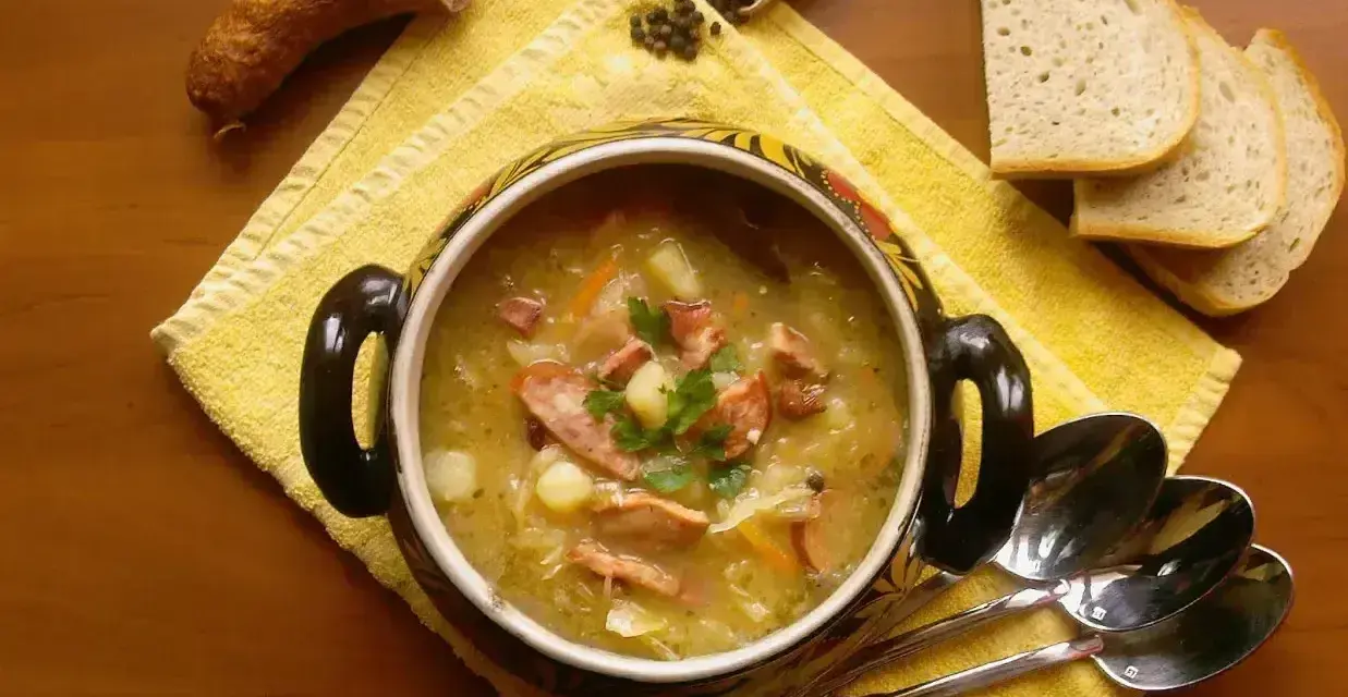 Aromaticzny kapuśniak jak u babci, z kiełbasą i ziemniakami, podany w ozdobnym garnku, obok kromek chleba i łyżek.