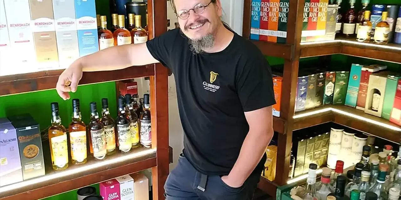 Mann in einem Guinness-T-Shirt steht in einem Laden voller Whisky-Flaschen und -Schachteln. Er lächelt und lehnt sich an ein Regal.