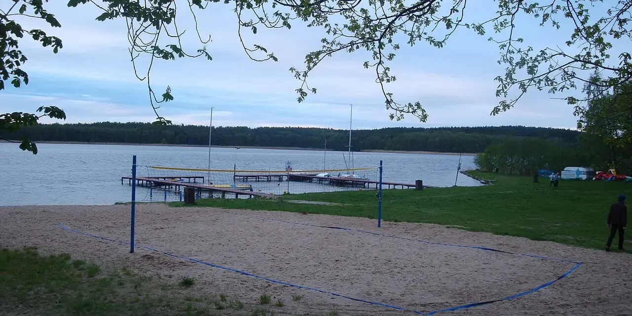 Plaża z boiskiem do siatkówki plażowej nad jeziorem w Borneńskim Sulimowie. W tle pomosty, żaglówki i las.